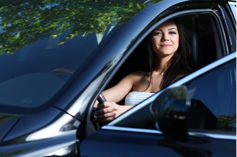 A woman sitting happily in her car. Getting the car insured is a vital expense that will protect you and relieve a big burden in case you get in an accident.