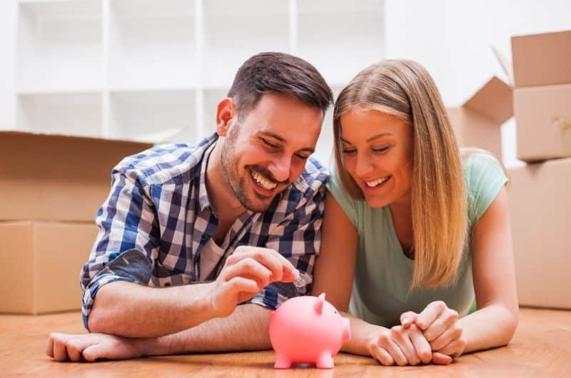 A young married couple is laying on the floor and putting money into a piggy bank. They're saving money towards a future house together.