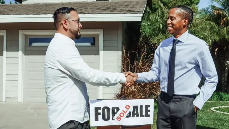 A realtor has just closed on a home for his recent client, they're shaking hands outside the house "sold" sign.