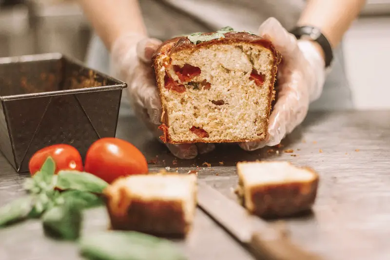 A baker is showing off a tomato basil loaf of bread that just came out of the oven. This baker makes money by shipping these loaves to customers around the country.